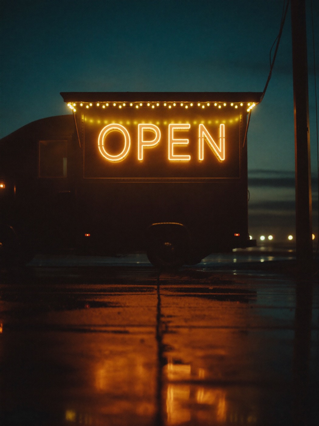 A glowing neon OPEN sign on a food truck reflected on wet pavement